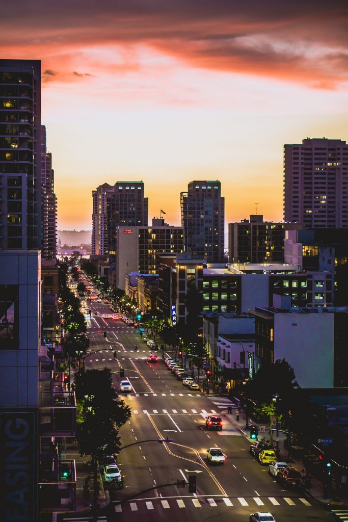 This image shows a view of San Diego's downtown. A street is pictured in the center of the image with tall buildings on either side. The photo was taken in the early evening.
