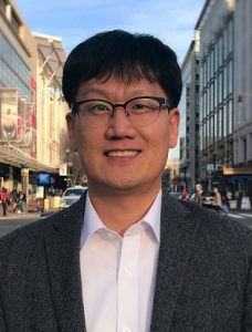 Portrait of Kyeongsu Kim standing outdoors while wearing a gray blazer with a white collared shirt.