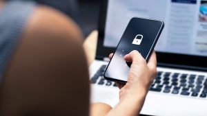 A smartphone user holds their phone with a lock icon displayed on the screen while working at a laptop computer.