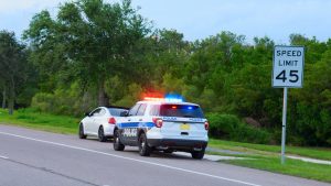 A police vehicle with flashing lights is parked behind a white sedan with a speed limit sign in the foreground.