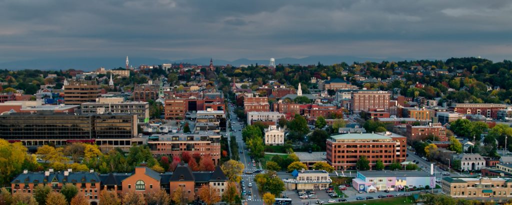 Aerial shot of Burlington, Vermont on a hazy evening in early Fall, looking along streets of historic buildings, shaded by trees that are beginning to to turn red, yellow and orange. The Green Mountains can be seen in the distance.