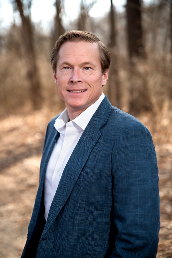 Smiling man standing in a wooded area with a suit jacket and shirt on.