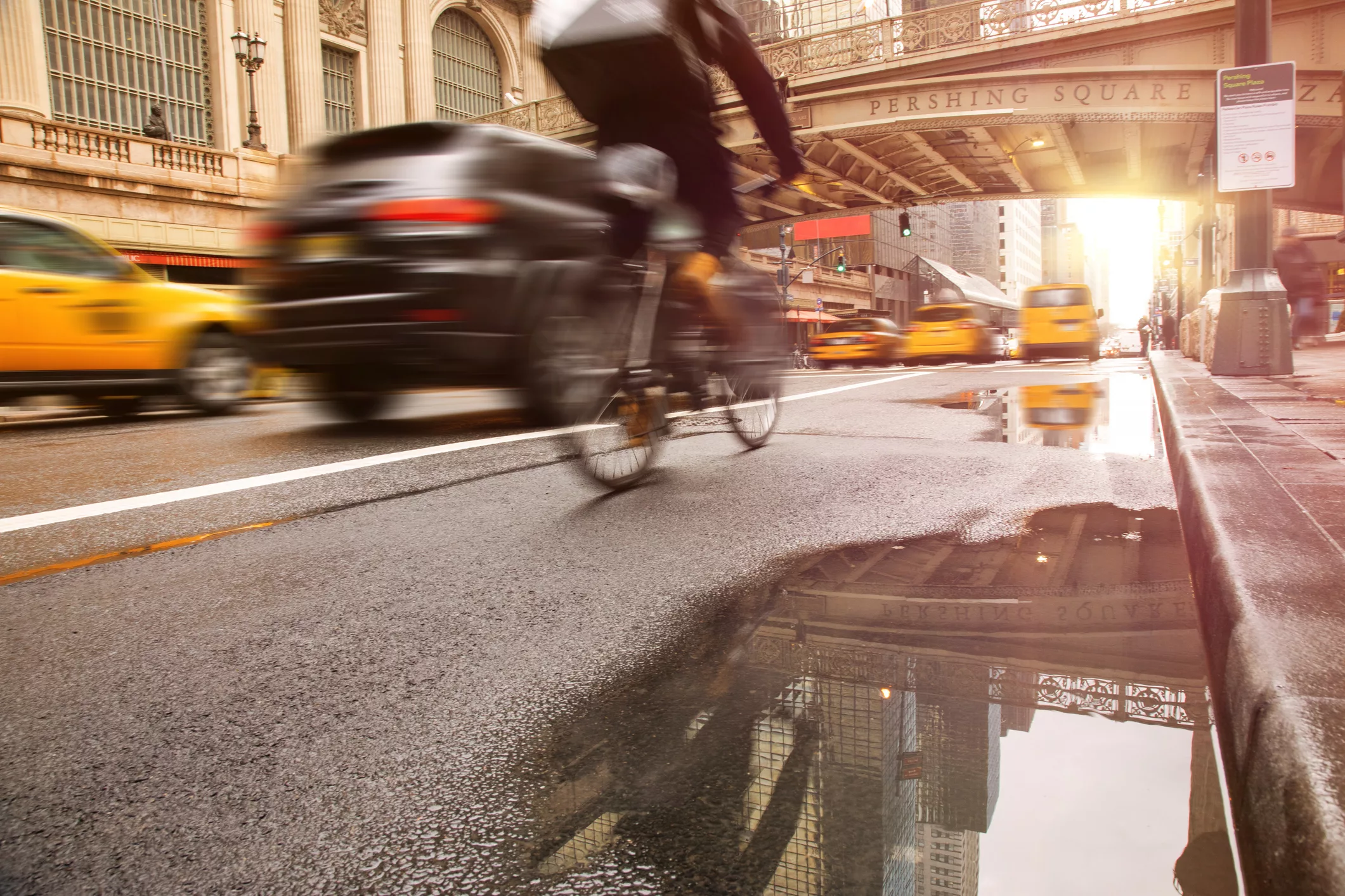 An urban street scene in motion: a cyclist rides alongside cars and yellow taxis on a wet road, with the Pershing Square bridge visible overhead and buildings reflected in a puddle. The image reflects the complexity of daily travel behavior that RSG’s AI-enabled surveys help classify and interpret.