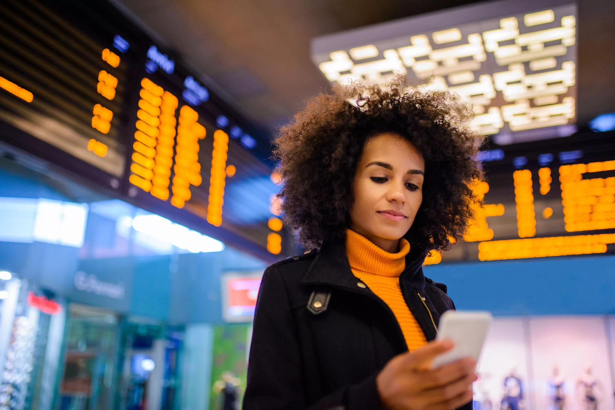 A woman at a transit hub checks her phone while transit departure boards glow in the background, representing real-world travel choices captured through modern survey tools.
