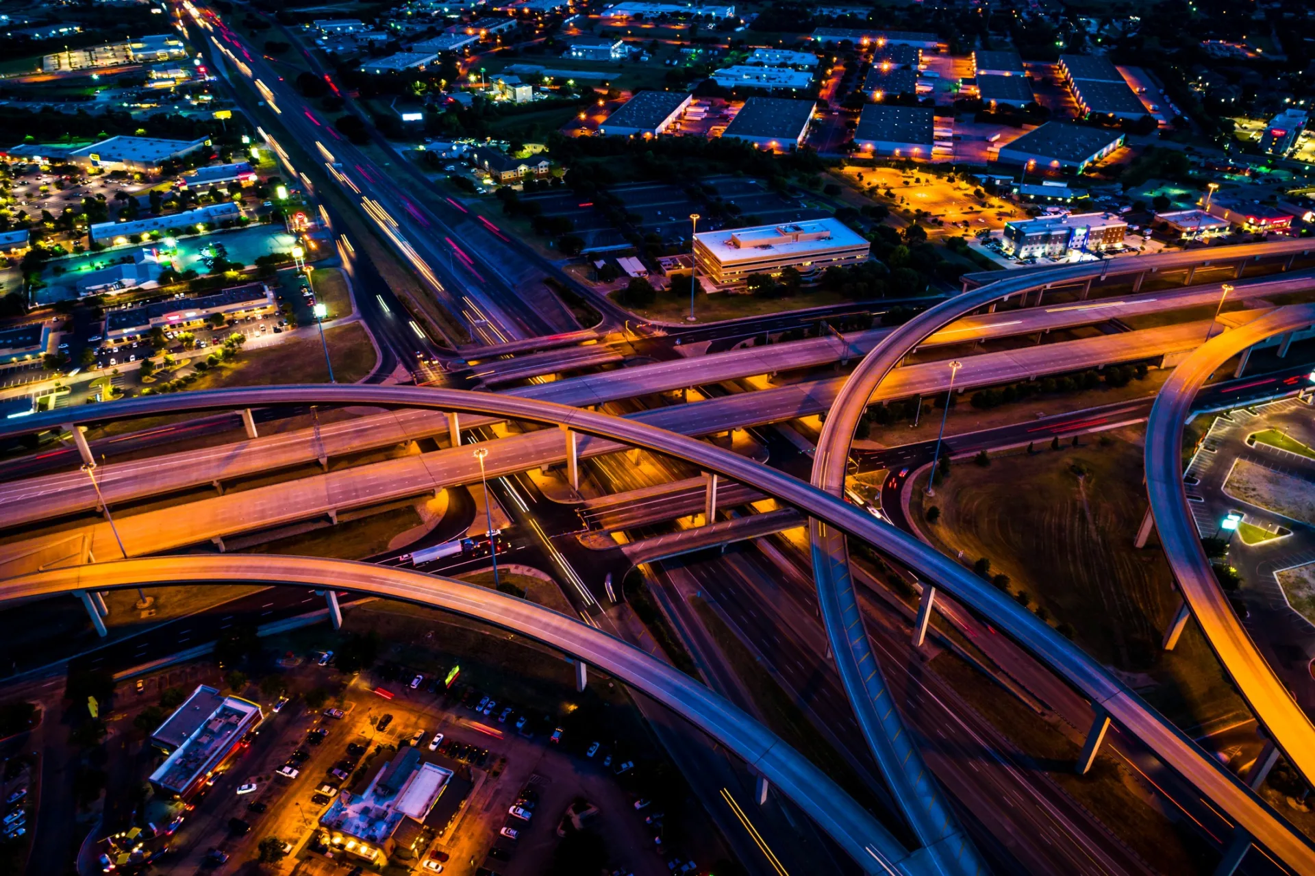 Aerial nighttime view of a multilevel highway interchange surrounded by commercial development and illuminated roadways.