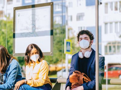 This image shows three people wearing masks while seated at an outdoors bus station during the day. Behind them is a map displaying the bus route.